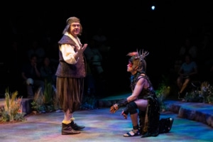 A performer in Shakespearean garb stands and gestures while addressing another performer in a fantastical costume who kneels on the stage and looks upward.