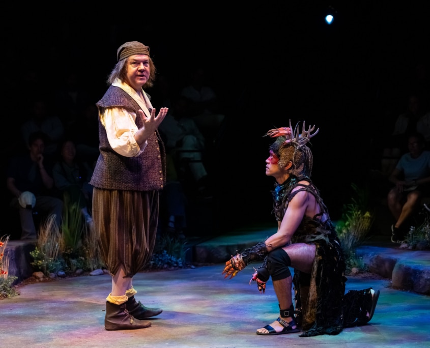 A performer in Shakespearean garb stands and gestures while addressing another performer in a fantastical costume who kneels on the stage and looks upward.