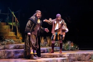 Two performers in Shakespearean costumes stand on stone steps in conversation on a stage decorated with grasses and wildflowers.
