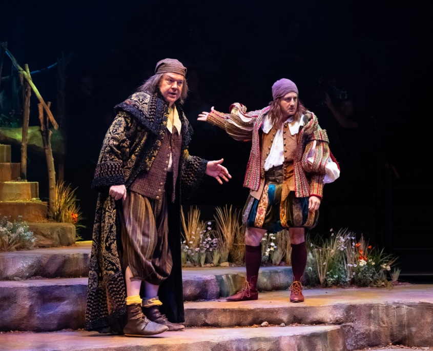 Two performers in Shakespearean costumes stand on stone steps in conversation on a stage decorated with grasses and wildflowers.
