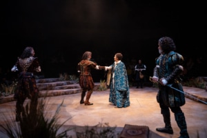 Two performances in Shakespearean garb hold each other’s outstretched arms while surrounded by other performers who stand ready to draw their swords.