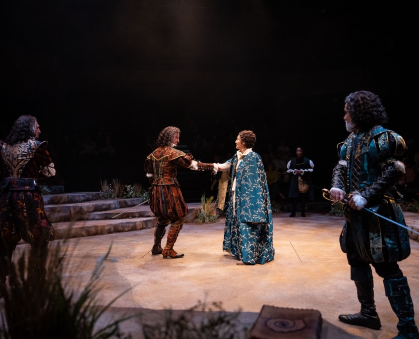 Two performances in Shakespearean garb hold each other’s outstretched arms while surrounded by other performers who stand ready to draw their swords.