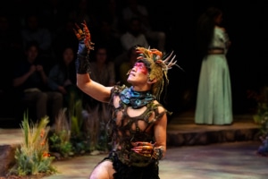 A performer in a fantasy-inspired costume kneels and raises a clawed hand toward the light.