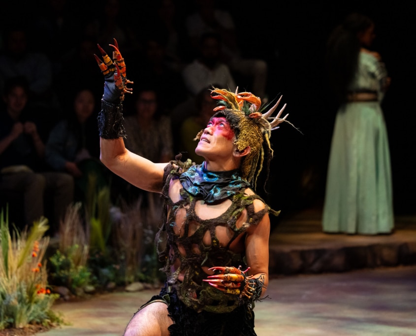 A performer in a fantasy-inspired costume kneels and raises a clawed hand toward the light.