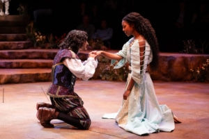 Two performers share a tender moment onstage as one kneels and kisses the other's hand. The pair wear richly detailed costumes and are surrounded by a circular stage set with stone terraces, flowers, and audience seating.