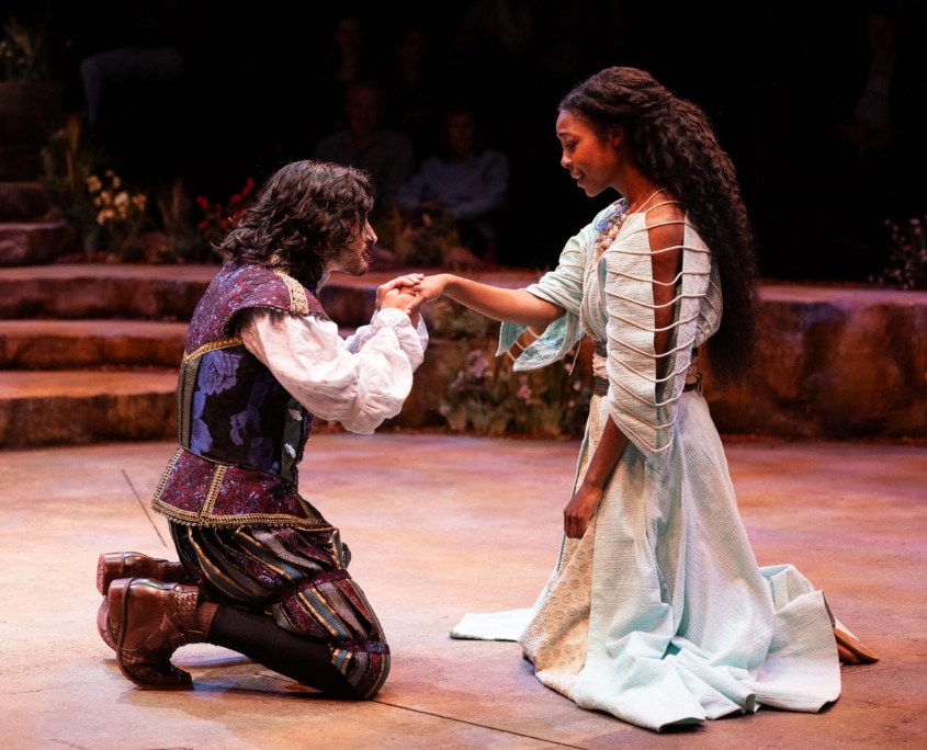 Two performers share a tender moment onstage as one kneels and kisses the other's hand. The pair wear richly detailed costumes and are surrounded by a circular stage set with stone terraces, flowers, and audience seating.