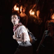 Actor in worn work clothes and a bandana stands in a dramatic spotlight while ensemble members in the background hold lanterns on a dimly lit stage.