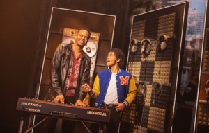 Two performers stand at a keyboard synthesizer on a studio‑themed set, surrounded by soundproof panels, headphones, and audio equipment.