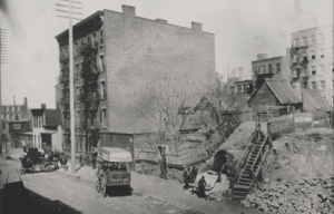 Historic photograph of a tenement‑era neighborhood with dirt roads and early urban structures