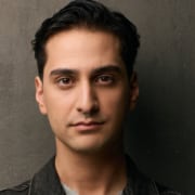 Person with short dark hair wearing a denim jacket against a textured wall