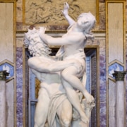 Marble sculpture depicting a powerful figure lifting another figure in a dynamic, twisting pose, displayed in an ornate gallery with marble columns, gilded architectural details, and a carved relief above the statue.