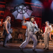 Performer in a light suit leads a lively dance while holding a large decorative umbrella, surrounded by ensemble members in mid-movement.