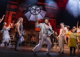 Performer in a light suit leads a lively dance while holding a large decorative umbrella, surrounded by ensemble members in mid-movement.