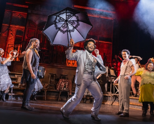 Performer in a light suit leads a lively dance while holding a large decorative umbrella, surrounded by ensemble members in mid-movement.