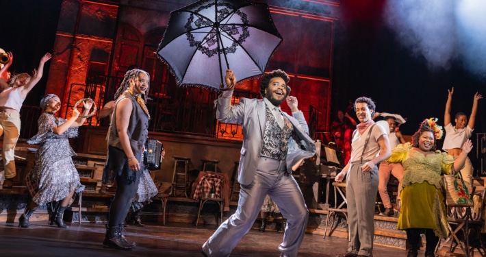 Performer in a light suit leads a lively dance while holding a large decorative umbrella, surrounded by ensemble members in mid-movement.