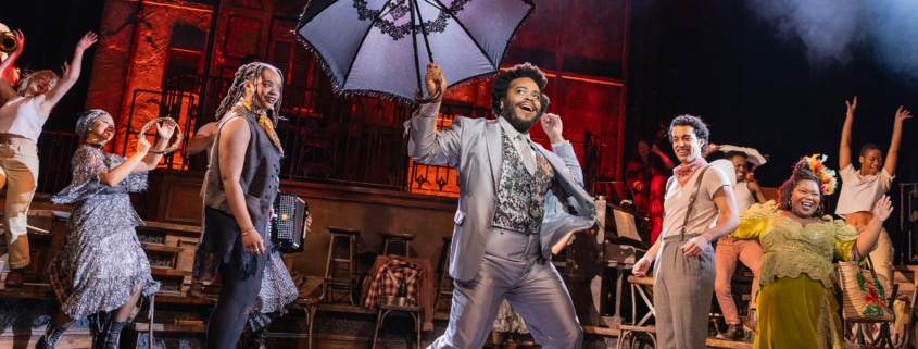 Performer in a light suit leads a lively dance while holding a large decorative umbrella, surrounded by ensemble members in mid-movement.