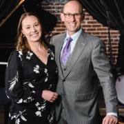 Two people standing side by side in a performance venue, wearing formal attire, with a grand piano and brick wall visible in the background.