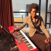Person seated at an electronic keyboard indoors, wearing a light-colored blazer and dark clothing, with a microphone stand nearby and large windows behind the scene.