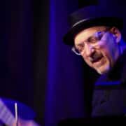 Person wearing a dark hat and glasses, seated at a piano under stage lighting, photographed from a slight angle during a live performance setting.