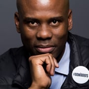 Close-up portrait of a person resting their chin on one hand, wearing a dark jacket with a circular button that includes visible text.