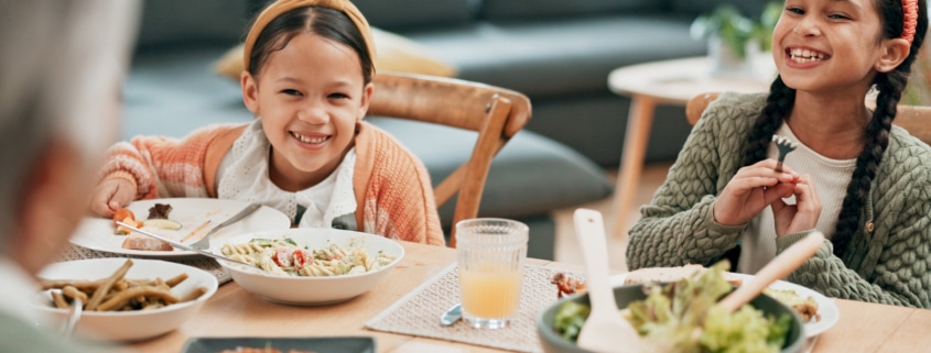 A dining table set with various dishes while two children sit and eat, with an older person seated in the foreground in a home living room setting.