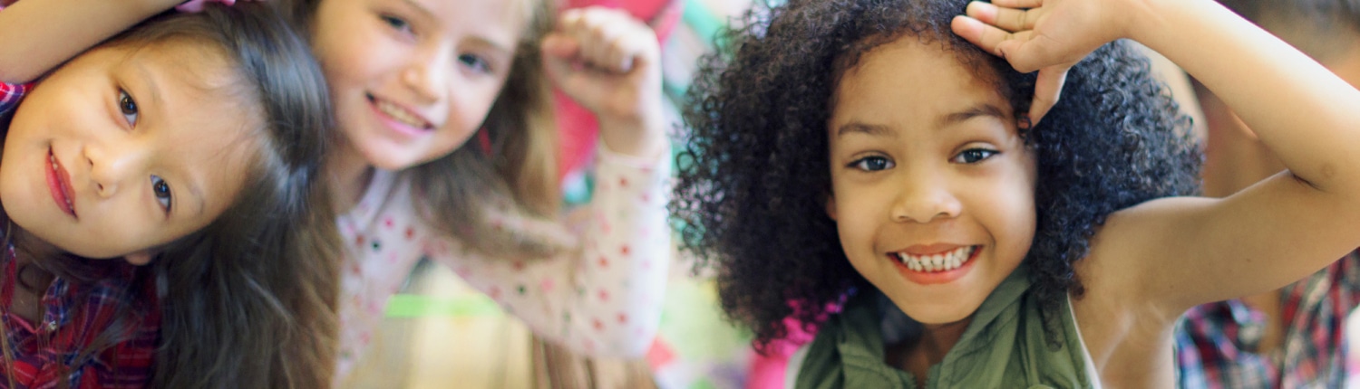 A group of children sitting closely together indoors, raising their arms and leaning toward the camera in a playful, energetic pose.