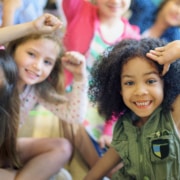 A group of children sitting closely together indoors, raising their arms and leaning toward the camera in a playful, energetic pose.