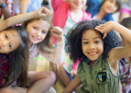 A group of children sitting closely together indoors, raising their arms and leaning toward the camera in a playful, energetic pose.