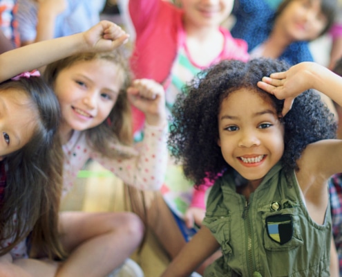 A group of children sitting closely together indoors, raising their arms and leaning toward the camera in a playful, energetic pose.