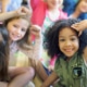 A group of children sitting closely together indoors, raising their arms and leaning toward the camera in a playful, energetic pose.