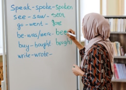 A person wearing a patterned outfit and a headscarf writes English irregular verbs on a whiteboard in a classroom. The whiteboard lists verbs such as “speak–spoke–spoken,” “see–saw–seen,” “go–went–gone,” “be–was/were–been,” “buy–bought–bought,” and “write–wrote–…”. Bookshelves are visible in the background.
