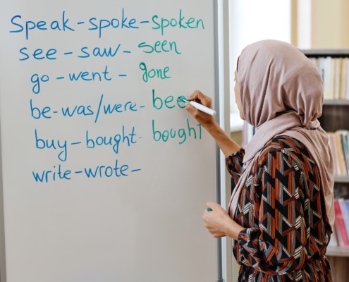 A person wearing a patterned outfit and a headscarf writes English irregular verbs on a whiteboard in a classroom. The whiteboard lists verbs such as “speak–spoke–spoken,” “see–saw–seen,” “go–went–gone,” “be–was/were–been,” “buy–bought–bought,” and “write–wrote–…”. Bookshelves are visible in the background.
