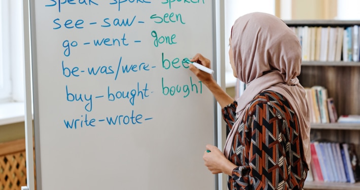 A person wearing a patterned outfit and a headscarf writes English irregular verbs on a whiteboard in a classroom. The whiteboard lists verbs such as “speak–spoke–spoken,” “see–saw–seen,” “go–went–gone,” “be–was/were–been,” “buy–bought–bought,” and “write–wrote–…”. Bookshelves are visible in the background.
