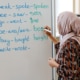 A person wearing a patterned outfit and a headscarf writes English irregular verbs on a whiteboard in a classroom. The whiteboard lists verbs such as “speak–spoke–spoken,” “see–saw–seen,” “go–went–gone,” “be–was/were–been,” “buy–bought–bought,” and “write–wrote–…”. Bookshelves are visible in the background.