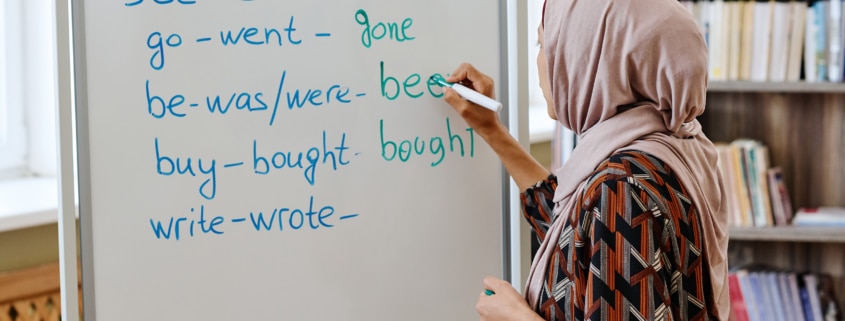 A person wearing a patterned outfit and a headscarf writes English irregular verbs on a whiteboard in a classroom. The whiteboard lists verbs such as “speak–spoke–spoken,” “see–saw–seen,” “go–went–gone,” “be–was/were–been,” “buy–bought–bought,” and “write–wrote–…”. Bookshelves are visible in the background.