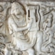 Ancient marble relief depicting a seated figure playing a lyre, surrounded by carved animals and smaller human figures in a detailed mythological scene.