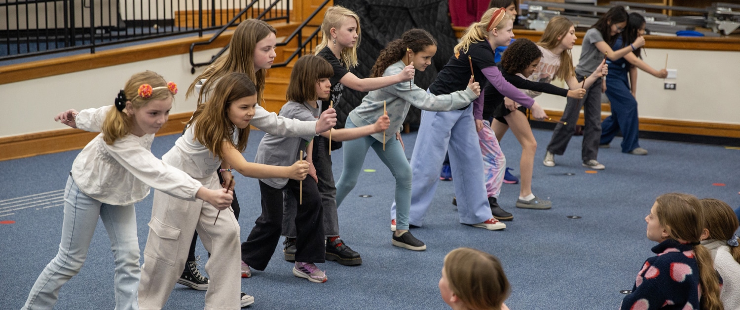 A line of elementary-age students practices staged combat movements with wooden sticks in a school auditorium during a Shakespeare combat workshop.
