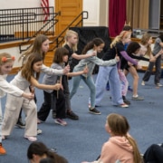 A line of elementary-age students practices staged combat movements with wooden sticks in a school auditorium during a Shakespeare combat workshop.
