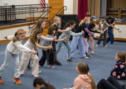 A line of elementary-age students practices staged combat movements with wooden sticks in a school auditorium during a Shakespeare combat workshop.