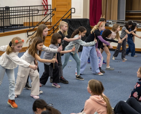 A line of elementary-age students practices staged combat movements with wooden sticks in a school auditorium during a Shakespeare combat workshop.