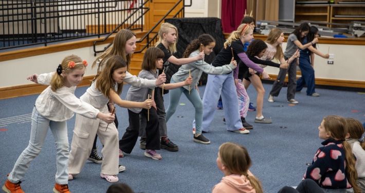 A line of elementary-age students practices staged combat movements with wooden sticks in a school auditorium during a Shakespeare combat workshop.