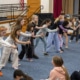 A line of elementary-age students practices staged combat movements with wooden sticks in a school auditorium during a Shakespeare combat workshop.
