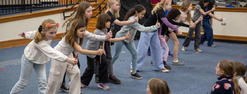 A line of elementary-age students practices staged combat movements with wooden sticks in a school auditorium during a Shakespeare combat workshop.