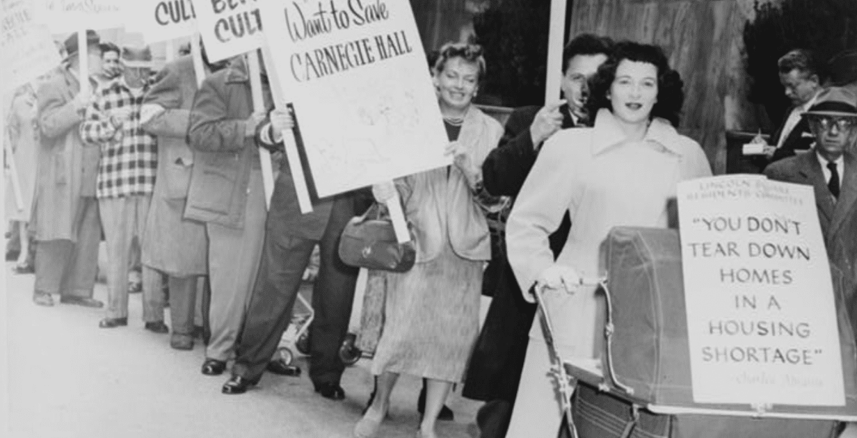 Black-and-white photo of a public protest outside a modern building, with people standing in a line holding signs that read ‘Shelter Before Culture’ and ‘You don’t tear down homes in a housing shortage,’ including one sign attached to a baby carriage.