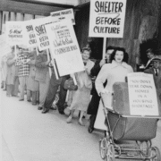 Black-and-white photo of a public protest outside a modern building, with people standing in a line holding signs that read ‘Shelter Before Culture’ and ‘You don’t tear down homes in a housing shortage,’ including one sign attached to a baby carriage.