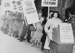 Black-and-white photo of a public protest outside a modern building, with people standing in a line holding signs that read ‘Shelter Before Culture’ and ‘You don’t tear down homes in a housing shortage,’ including one sign attached to a baby carriage.