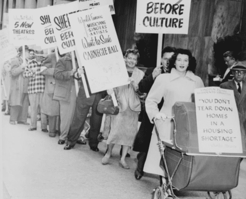 Black-and-white photo of a public protest outside a modern building, with people standing in a line holding signs that read ‘Shelter Before Culture’ and ‘You don’t tear down homes in a housing shortage,’ including one sign attached to a baby carriage.