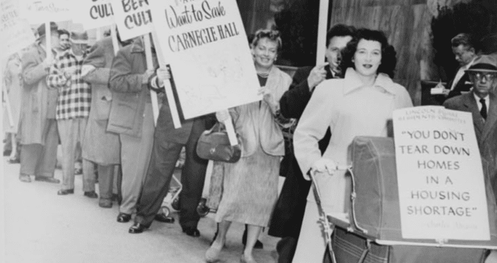 Black-and-white photo of a public protest outside a modern building, with people standing in a line holding signs that read ‘Shelter Before Culture’ and ‘You don’t tear down homes in a housing shortage,’ including one sign attached to a baby carriage.