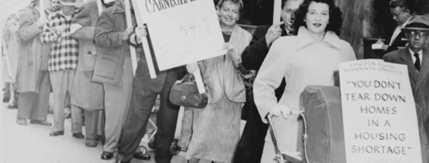 Black-and-white photo of a public protest outside a modern building, with people standing in a line holding signs that read ‘Shelter Before Culture’ and ‘You don’t tear down homes in a housing shortage,’ including one sign attached to a baby carriage.