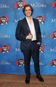 Person posing on a step-and-repeat backdrop featuring Les Misérables artwork, wearing a dark suit and white shirt, standing on a wooden floor at a promotional event.